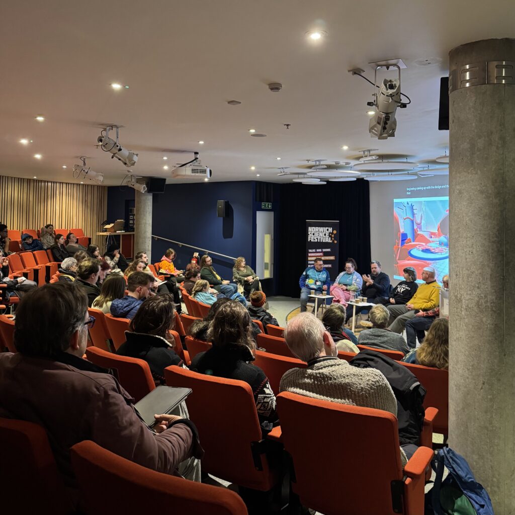 A group of people sit in red theater-style seats, facing a panel of speakers at the front of a modern, well-lit conference room. An education game presentation is displayed on the screen behind the panelists.