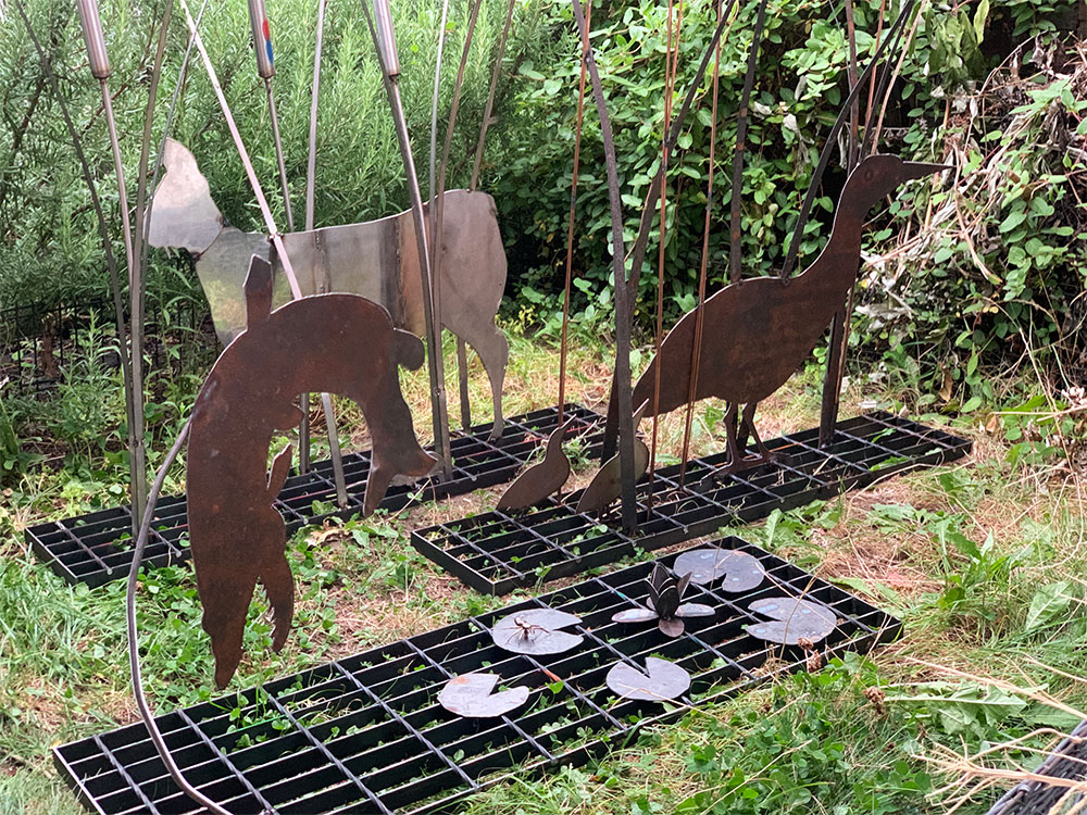 Metal silhouettes of animals stand among tall plants on black grates. Metal lily pads are on one of the grates, with green foliage in the background.