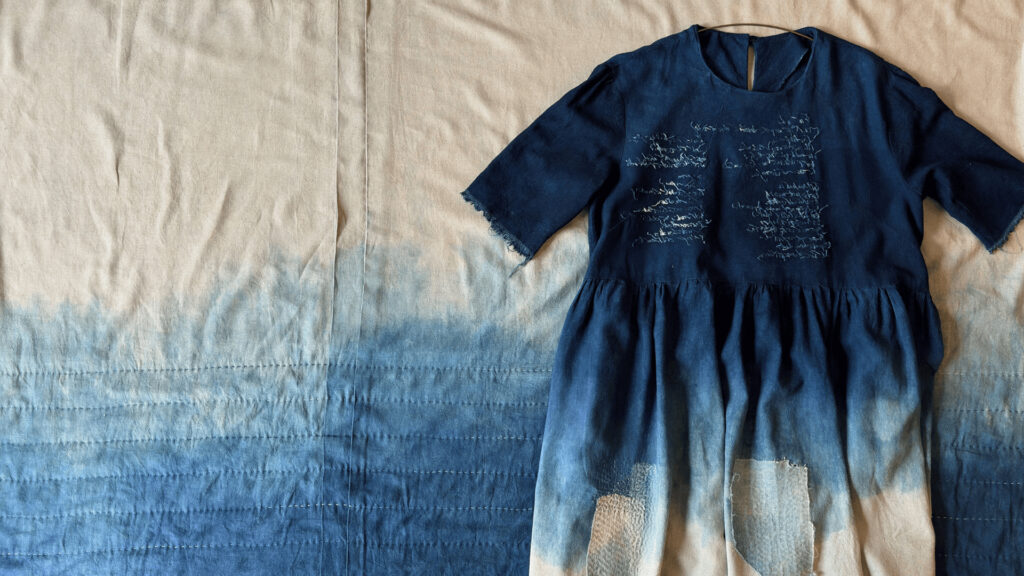 A blue and beige ombré dress with white embroidered writing lies on a matching ombré fabric background, evoking a sense of calm—perfect inspiration for your Online Open Day outfit. The short sleeves and gathered waist blend beautifully into the scene.