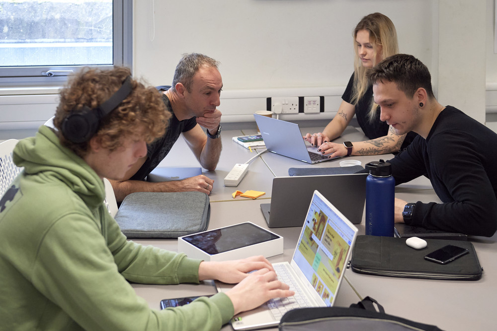 Four people sit around a table working on laptops in a bright room. One leans forward, observing a screen, while others focus on their work. Headphones, notebooks and a water bottle are on the table.