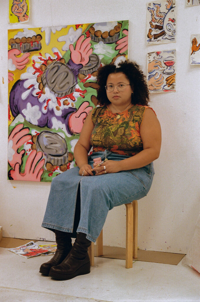 A person with curly hair, glasses, and a sleeveless patterned top sits on a stool in an art studio, surrounded by colorful abstract paintings and drawings with bold hand and face motifs.