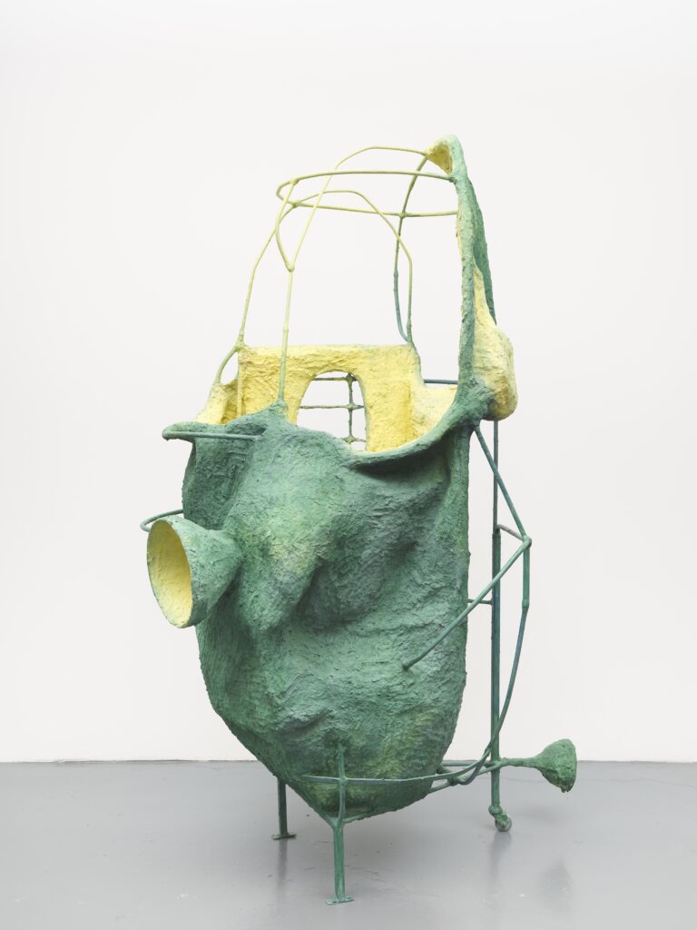 A large abstract sculpture with a textured green and yellow surface, featuring curved and angular forms supported by a thin metal frame, displayed against a plain white background.
