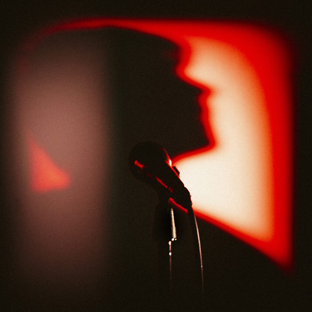 A microphone stands in focus in front of a red-lit background, where the blurry shadow of a person's head and shoulders is visible in profile.
