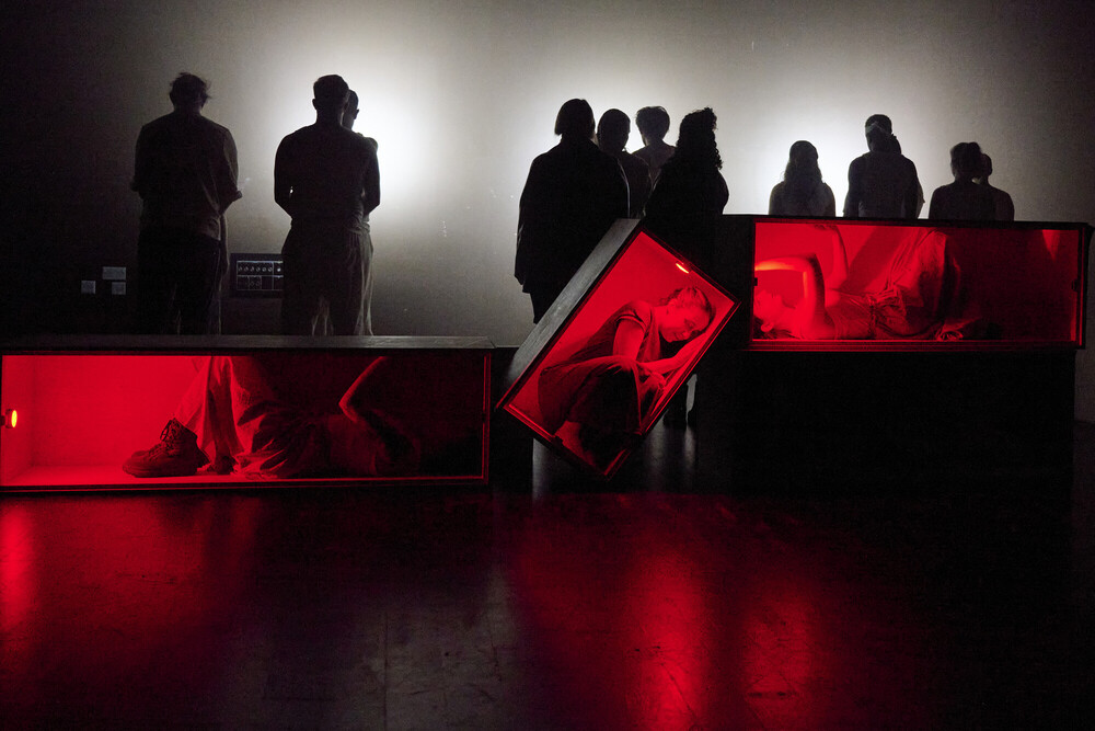Silhouetted figures stand in front of a bright light, while three people lie in transparent boxes illuminated with red light on the floor in the foreground, creating a dramatic and mysterious scene.