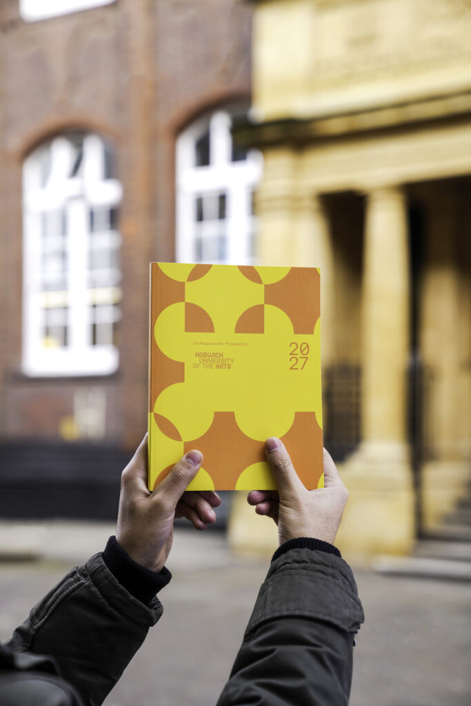 A person holds up a yellow and orange book titled "2027" with abstract shapes on the cover, standing outside near a building with large windows and stone columns.