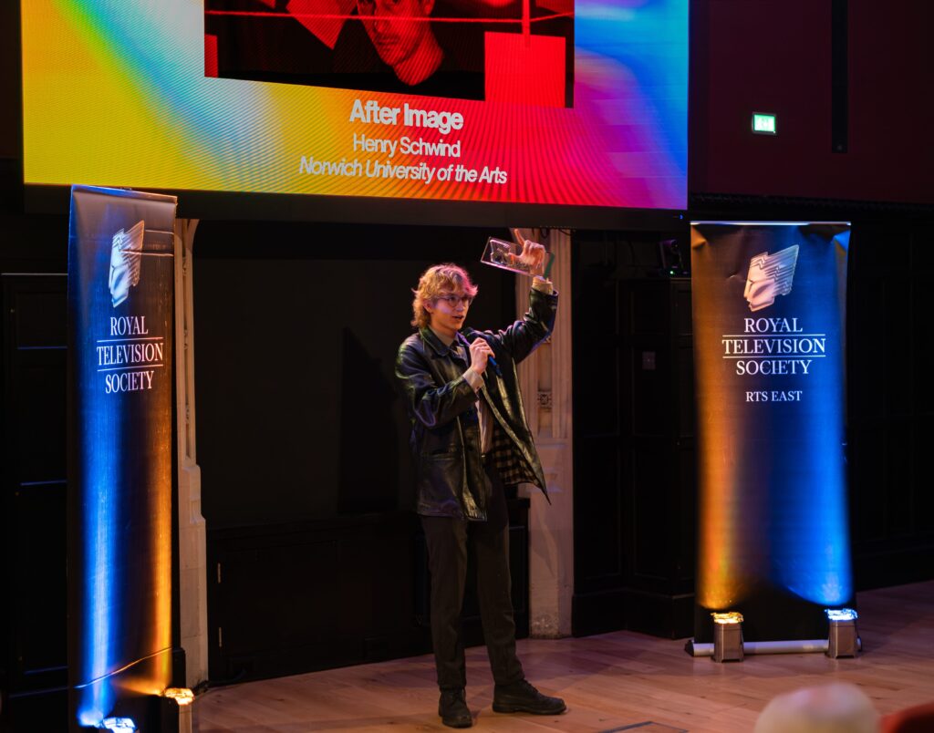 A person holds up a trophy and speaks into a microphone on stage at an awards event, with two Royal Television Society banners and a large screen displaying "After Image" and "Norwich University of the Arts" behind them.
