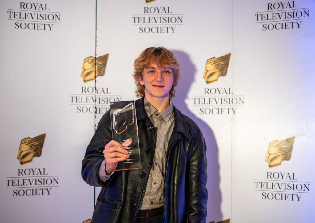 A person with blond hair in a leather jacket holds up an award and smiles in front of a Royal Television Society backdrop.