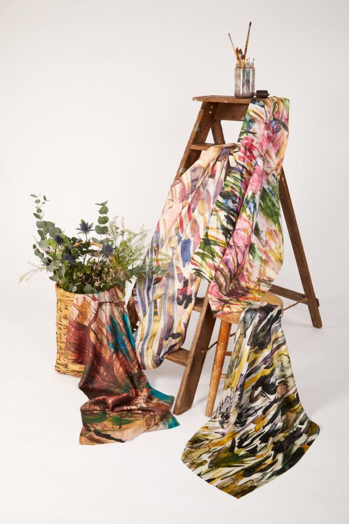 A wooden ladder displays colourful, abstract-patterned scarves draped over its steps. A glass jar with paintbrushes sits on top, and a woven basket with green foliage is placed beside the ladder on a plain white background.
