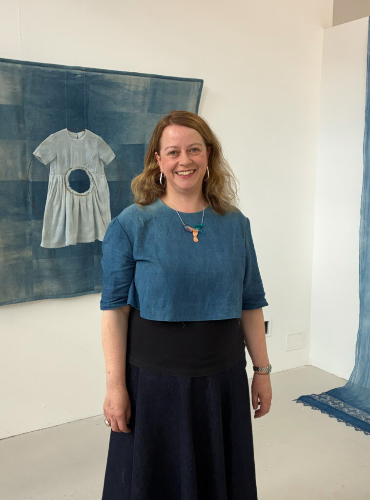 A woman with wavy, light brown hair stands smiling in an art gallery. She wears a blue top and skirt. Behind her, a blue artwork with a child’s dress is displayed on the wall.