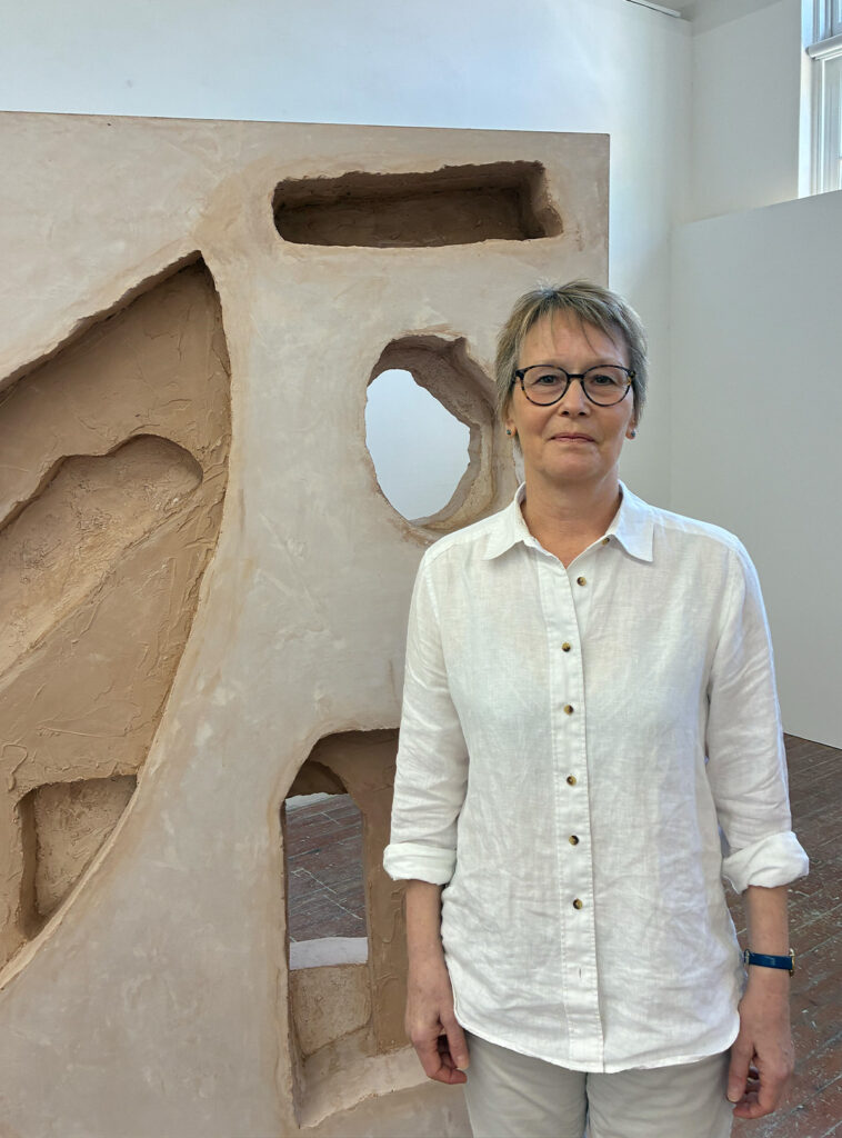 A person wearing glasses and a white button-up shirt stands indoors in front of a beige textured abstract sculpture with irregular cut-outs. The setting appears to be an art gallery or studio.