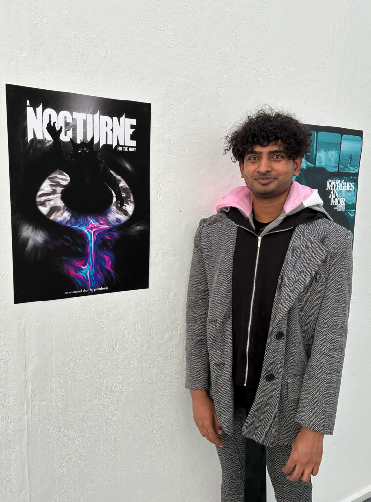 A person with curly hair stands beside a white wall displaying two posters. The closer poster reads "NOCTURNE" and features abstract, dark artwork with bright colors at the center. The person wears a grey coat over a hoodie.
