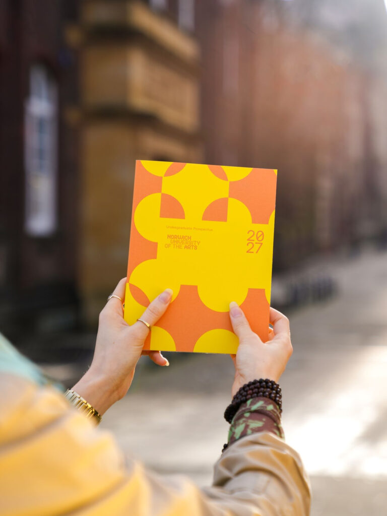 A person holds a colorful booklet with a bold yellow and orange geometric design. The cover reads "2027" and "Northern Festival of the Arts." The background shows a blurred street and buildings in soft daylight.