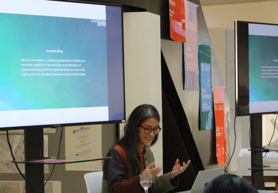 A woman with long gray hair and glasses speaks at a podium in front of two screens displaying a presentation. Colorful posters hang on the wall behind her. The text on the screen discusses “re-orienting.”.