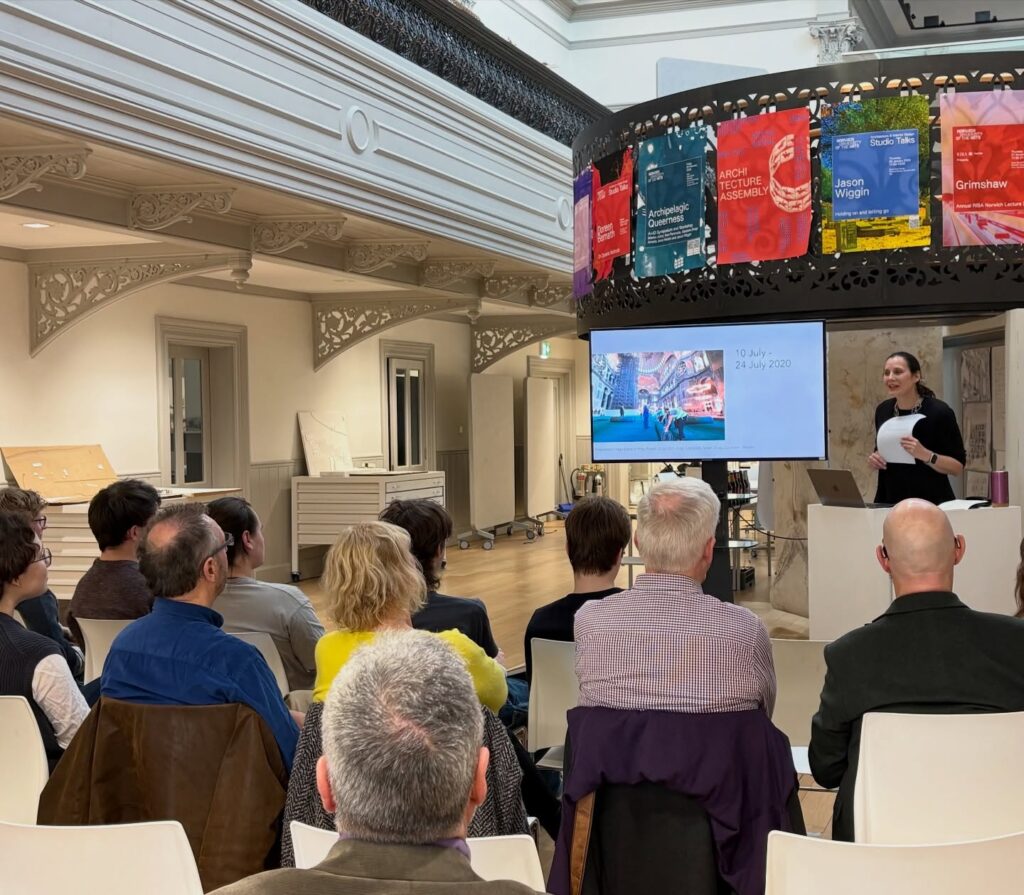 A woman stands at a podium giving a presentation to a seated audience in a modern room. Behind her, a screen displays slides and colorful posters hang on a balcony railing above.