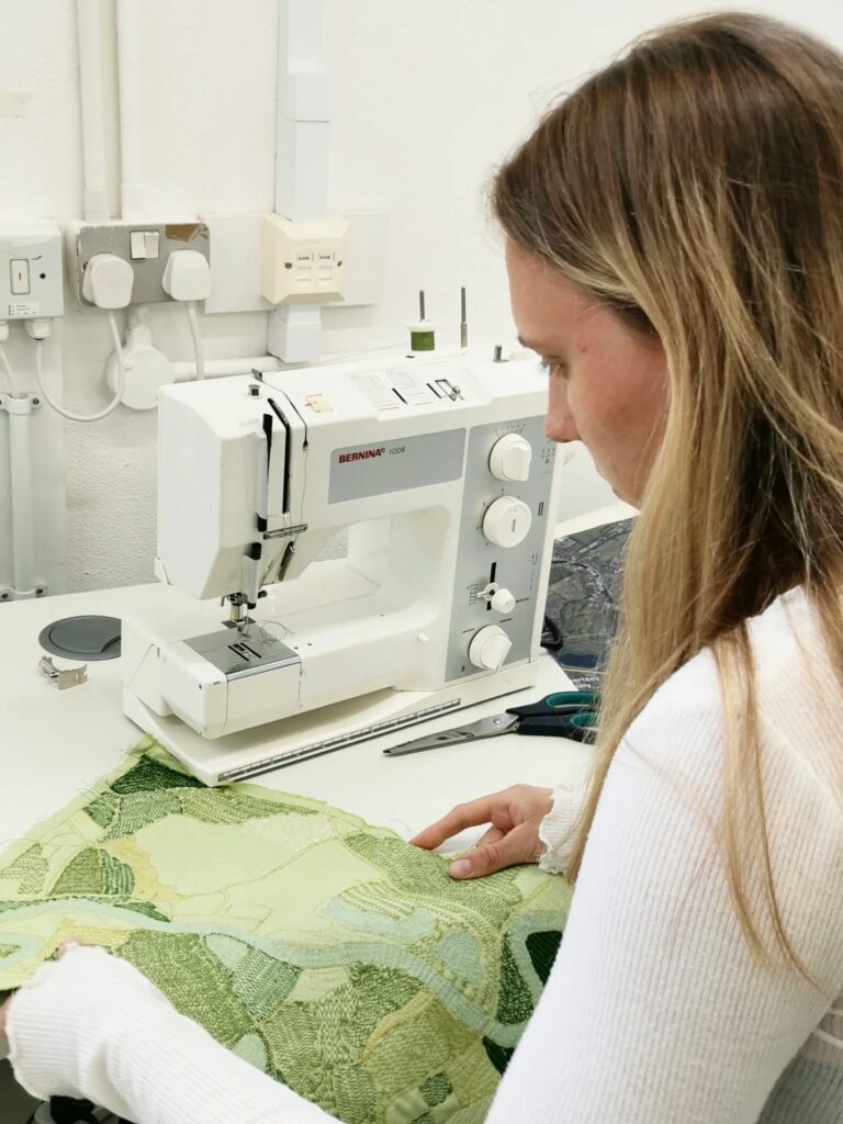 A woman with long blonde hair sits at a sewing machine, carefully working on a piece of green patterned fabric in a bright, modern workspace. Scissors and sewing tools are visible on the table.