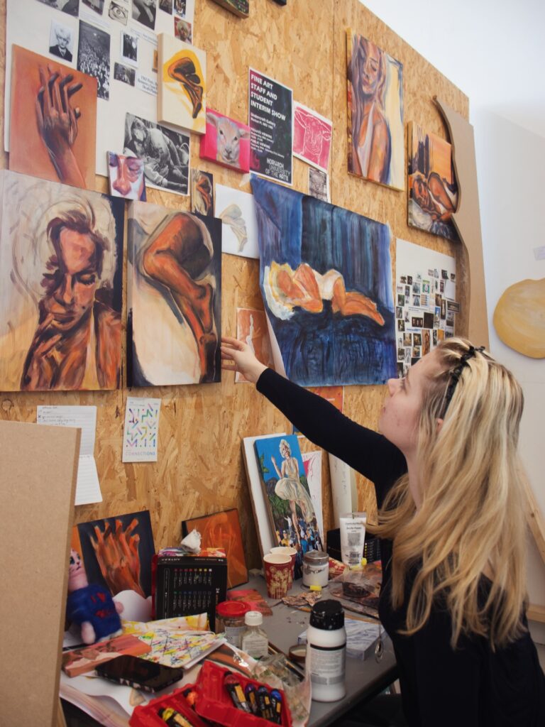 A woman with long blonde hair sits at a cluttered art desk, reaching up to touch a painting on a wall covered with various artworks, photographs, and sketches in a creative studio space.