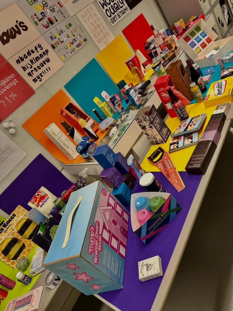 A display of various colorful products and packaging on tables, featuring cosmetics, food items, and gift boxes, with vibrant colored backgrounds and posters on the wall behind them.
