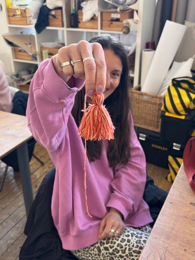 A person in a pink sweater holds up a handmade orange tassel, smiling. The background shows shelves with craft supplies and a wooden floor.