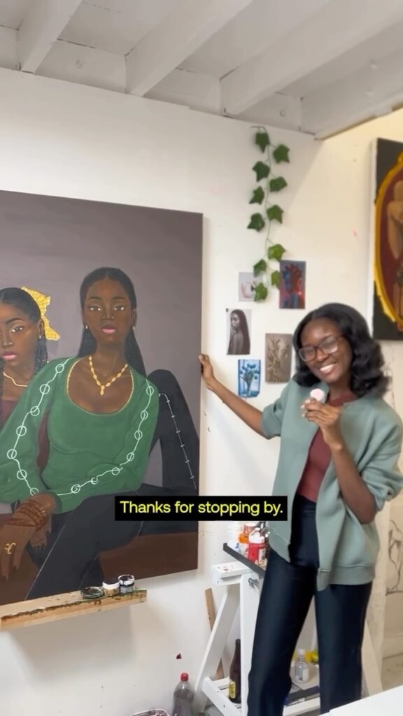 A smiling woman stands in an art studio holding a paintbrush, next to a large painting of two women. The wall behind her is decorated with artwork and greenery. The text on the image reads, "Thanks for stopping by.