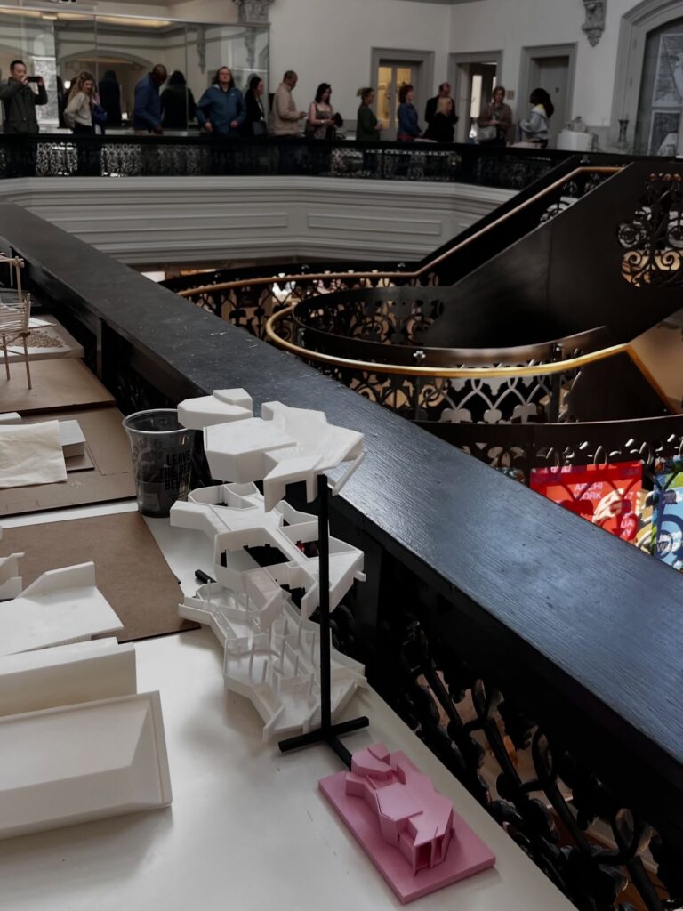 A display of small architectural models on a table near a decorative railing, with people standing and observing on the upper floor in a spacious building interior. A staircase and colorful stained glass are visible below.