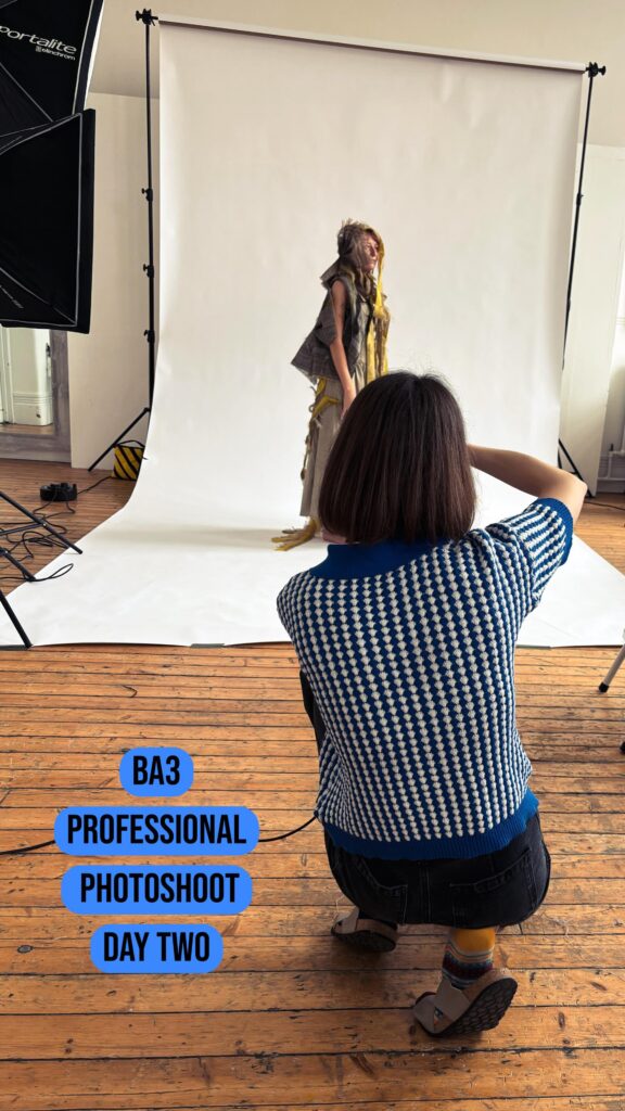 A photographer kneels on a wooden floor, taking photos of a model dressed in creative, layered clothing in front of a white backdrop in a studio. Text on the image reads: "BA3 Professional Photoshoot Day Two.