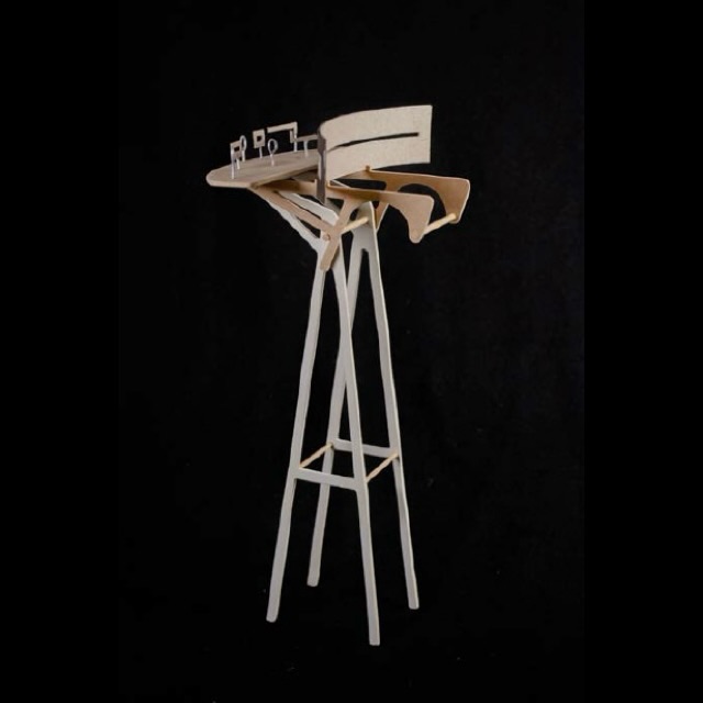 A sculptural wooden structure stands on tall, thin legs, with an abstract, layered top resembling a miniature architectural model, set against a plain black background.