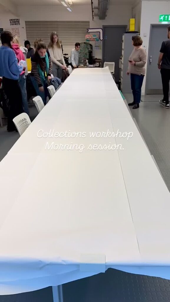 A group of people stands around a long table covered in white paper in a bright room. Text on the image reads, “Collections workshop Morning session.”.