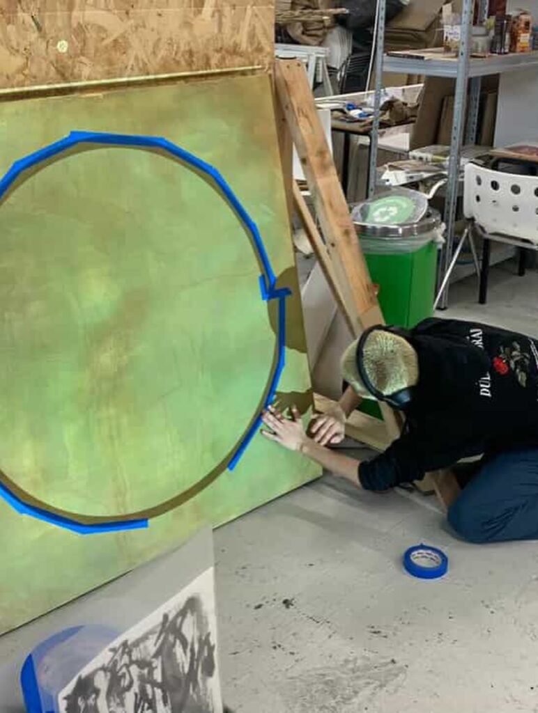 A person wearing a cap and black hoodie kneels on the floor, applying blue painter’s tape in a circular shape on a large, green-painted board in an art studio with shelves and supplies in the background.