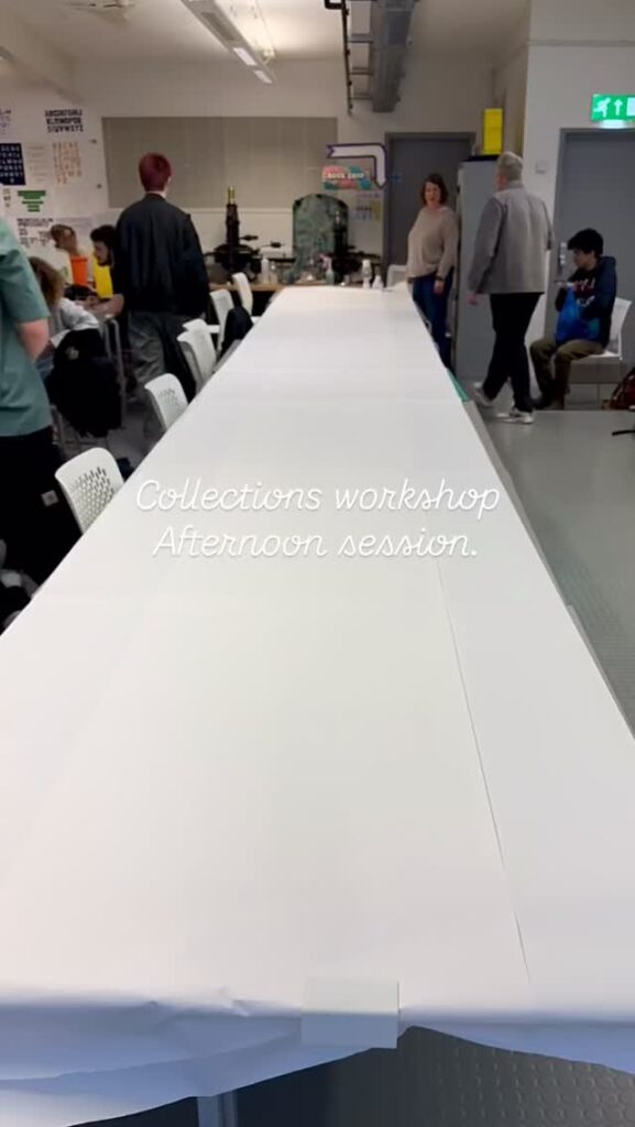 A long table covered with white paper is set up in a classroom with several people standing and sitting around. Text overlay reads, “Collections workshop Afternoon session.”.
