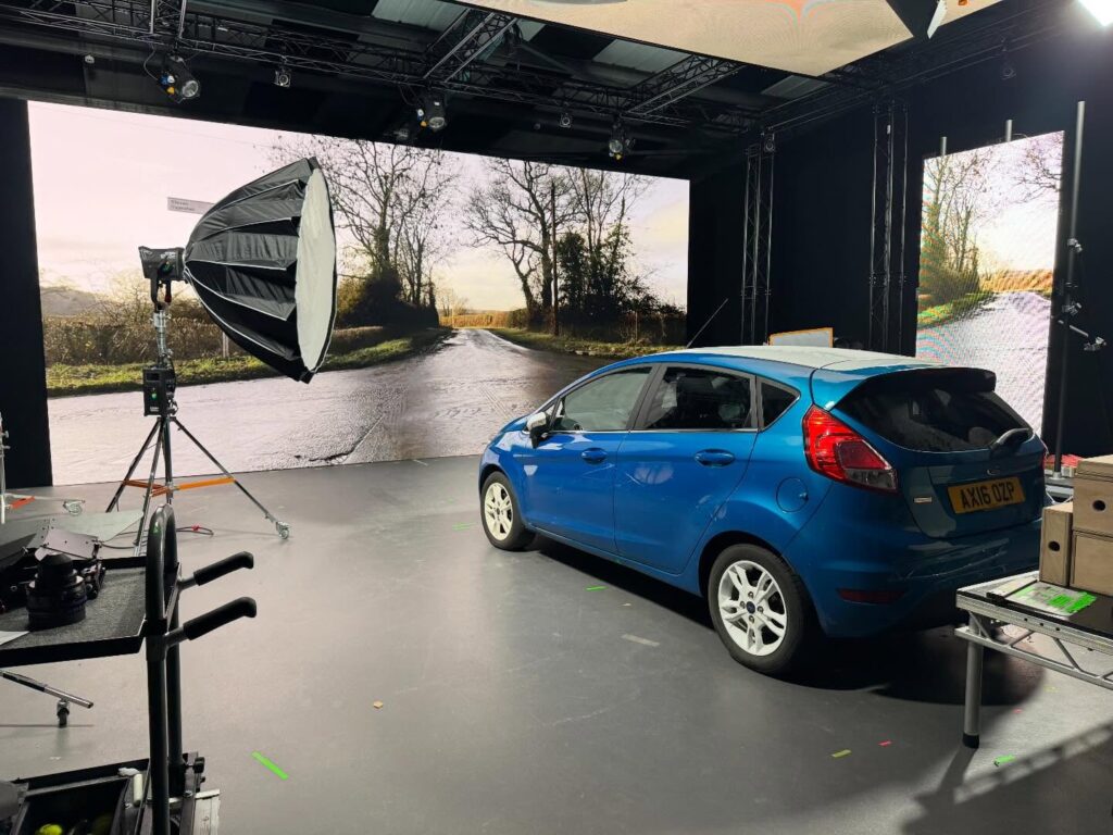 A blue car is parked inside a studio, surrounded by lighting equipment and film gear. A large screen behind the car displays a sunny rural road scene with trees and wet pavement.