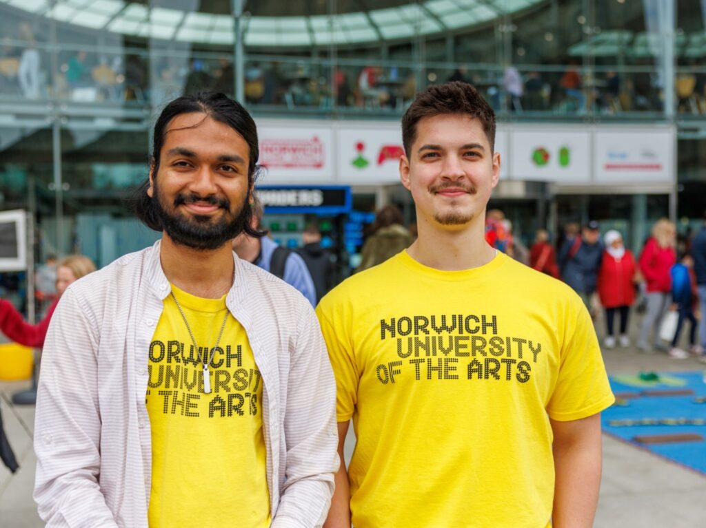 Two young men wearing matching yellow "Norwich University of the Arts" T-shirts stand side by side, smiling, in an outdoor area with a crowd and glass building in the background.
