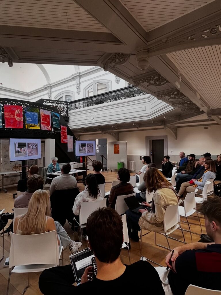 A group of people sit in white chairs facing a speaker at the front of a spacious, ornate room with high ceilings. The speaker presents slides on two large screens, and colorful posters hang above.