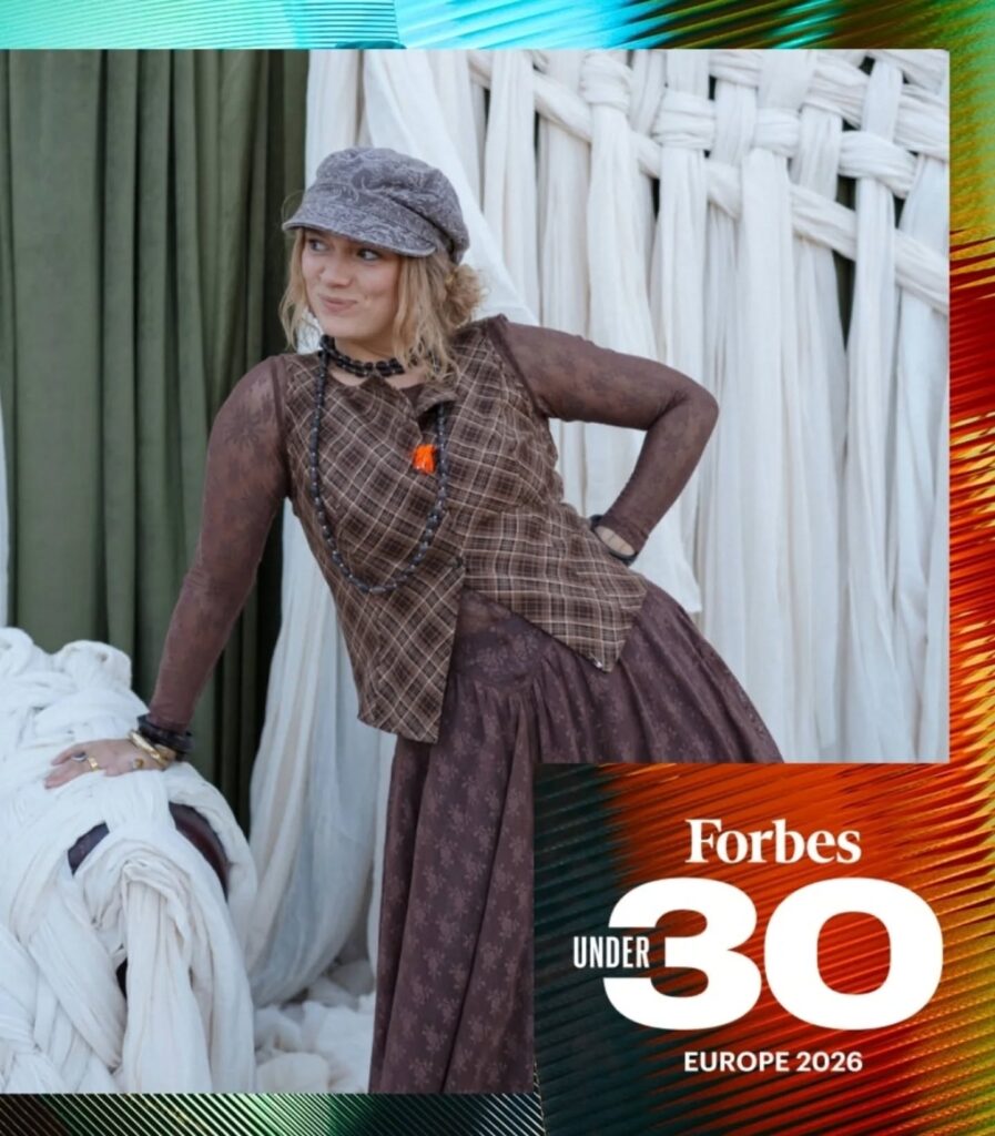 A smiling young woman in vintage-style clothing and a flat cap poses in front of large braided white fabric. The image has a Forbes Under 30 Europe 2026 graphic in the bottom right corner.