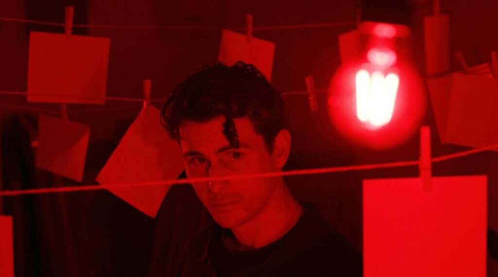A man stands in a dimly lit red room with blank sheets of paper hanging on lines behind him. A red light bulb glows brightly in the foreground, casting an intense red hue over the scene.
