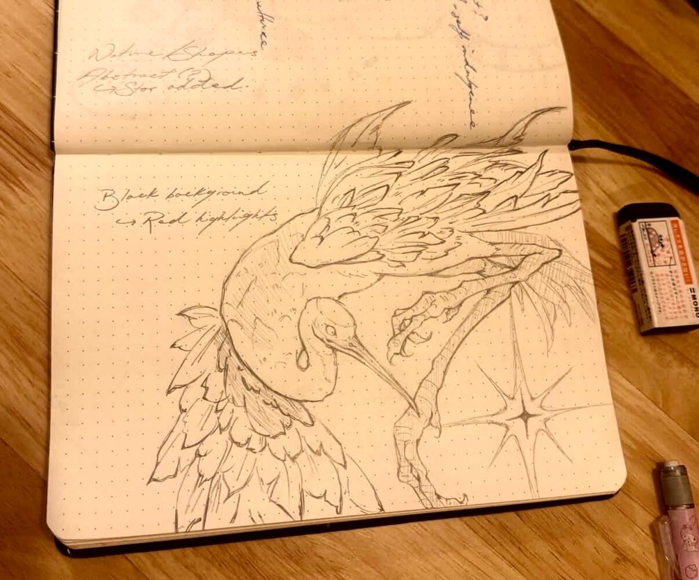 An open dotted notebook displays a detailed pencil sketch of a crane with outstretched wings and a star-like accent. Handwritten notes appear above the drawing, with an eraser and pen nearby on a wooden surface.