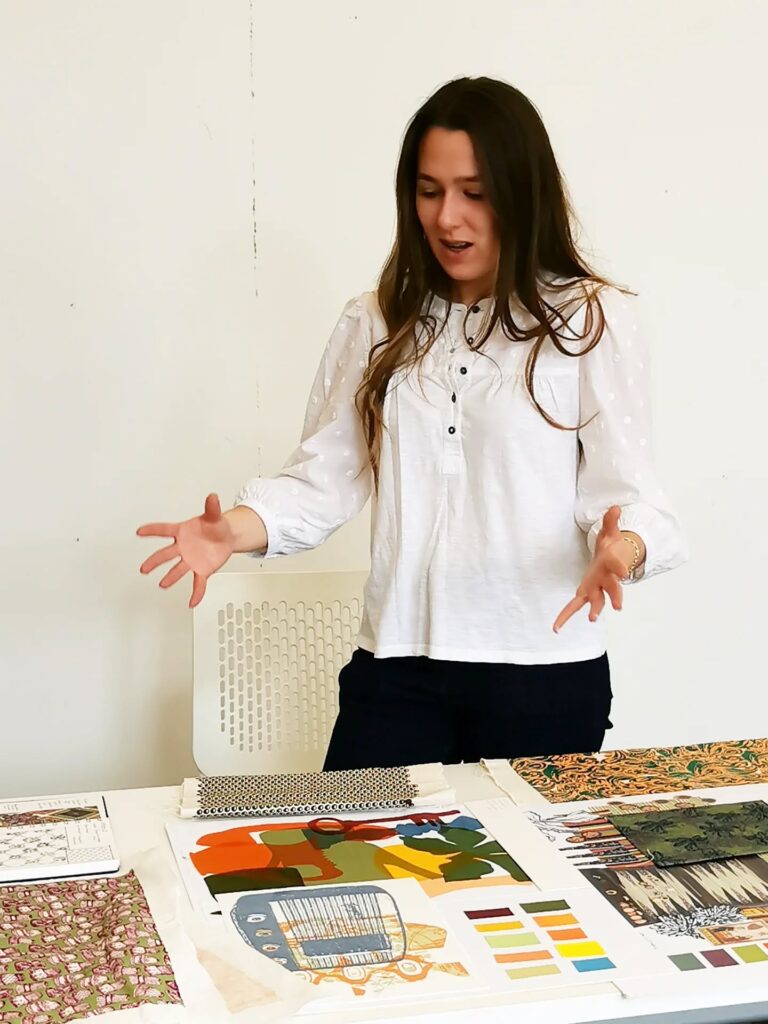 A woman with long brown hair, wearing a white blouse and dark pants, stands behind a table displaying colorful fabric samples and printed materials, gesturing expressively with both hands.