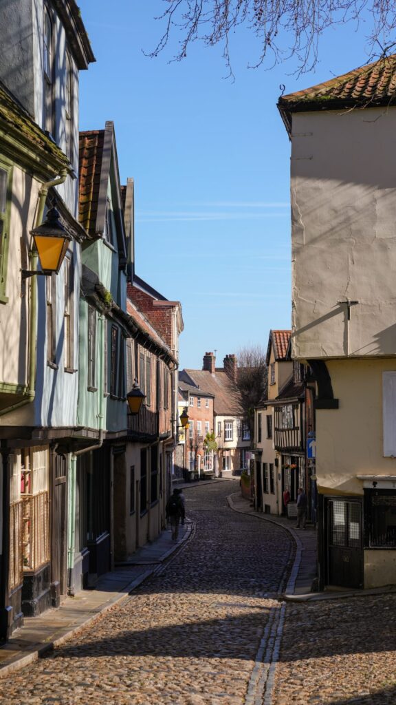 A narrow, cobblestone street curves between old, colorful buildings with overhanging upper floors, under a clear blue sky, in a quaint, historic town.