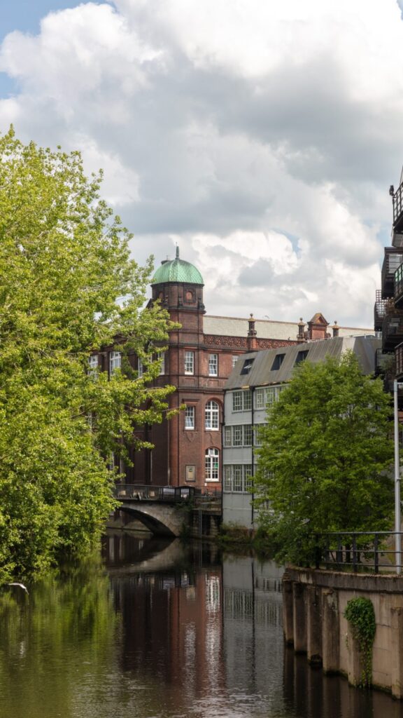 A historic brick building with a green dome stands beside a calm river, partly framed by leafy trees. The scene is reflected in the water under a partly cloudy sky.