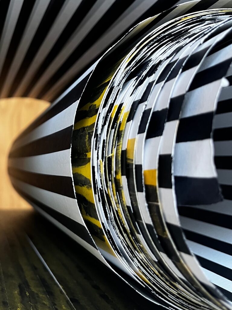 A close-up of rolled paper with black, white, and yellow stripes and checkered patterns, creating an abstract, geometric visual with a tunnel-like effect and strong lines.