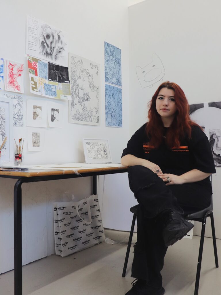 A person with long red hair sits on a chair beside a desk in an art studio, surrounded by various black-and-white and blue art pieces and sketches displayed on the walls. Art materials and a bag are on the floor.