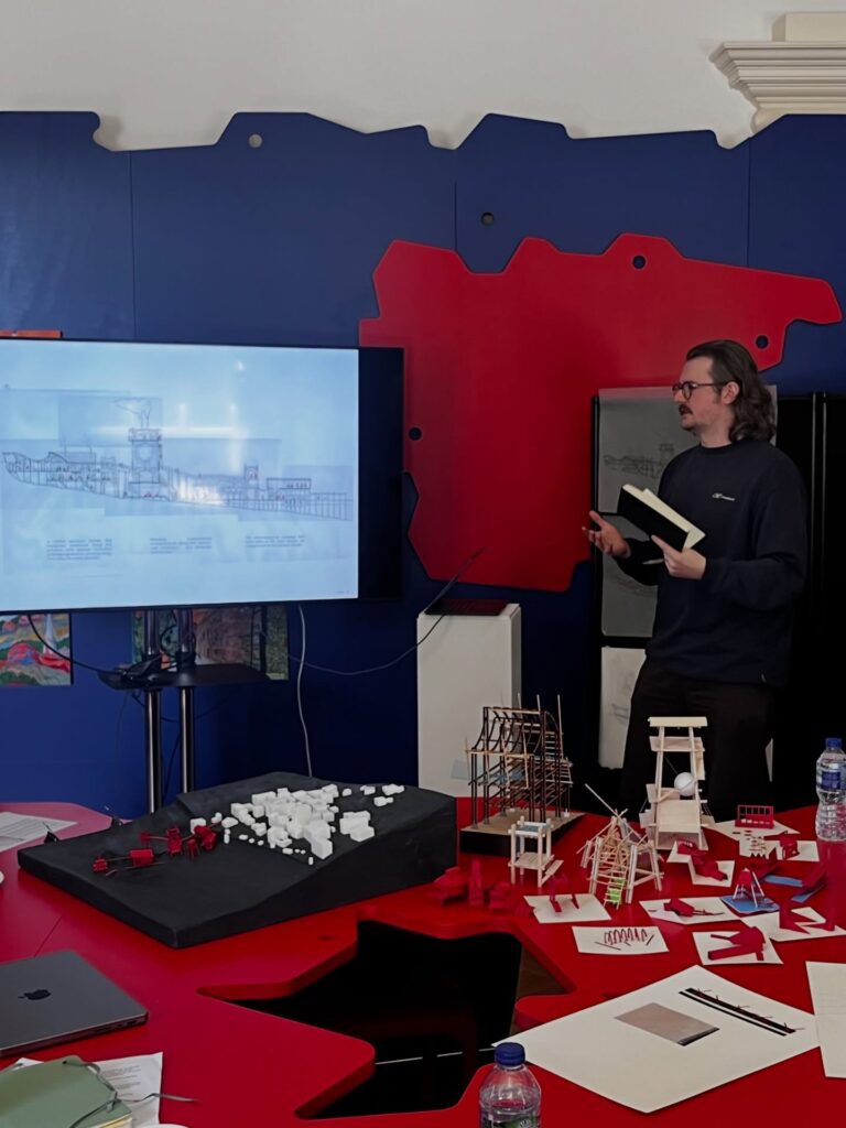 A man with glasses presents architectural designs on a screen in a colorful room. Table in front of him holds scale models, papers, and water bottles. A red and blue abstract wall decorates the background.