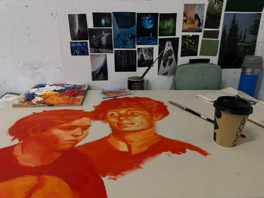 A partially painted portrait of two people on a table, surrounded by art supplies, a coffee cup, and a palette. The background wall is covered with various photos and art prints.