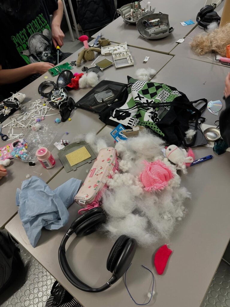 A cluttered table with fluffy stuffing, headphones, a pink pencil case, a patterned bag, stuffed animal parts, various craft supplies, and electronic components, with several people working around it.
