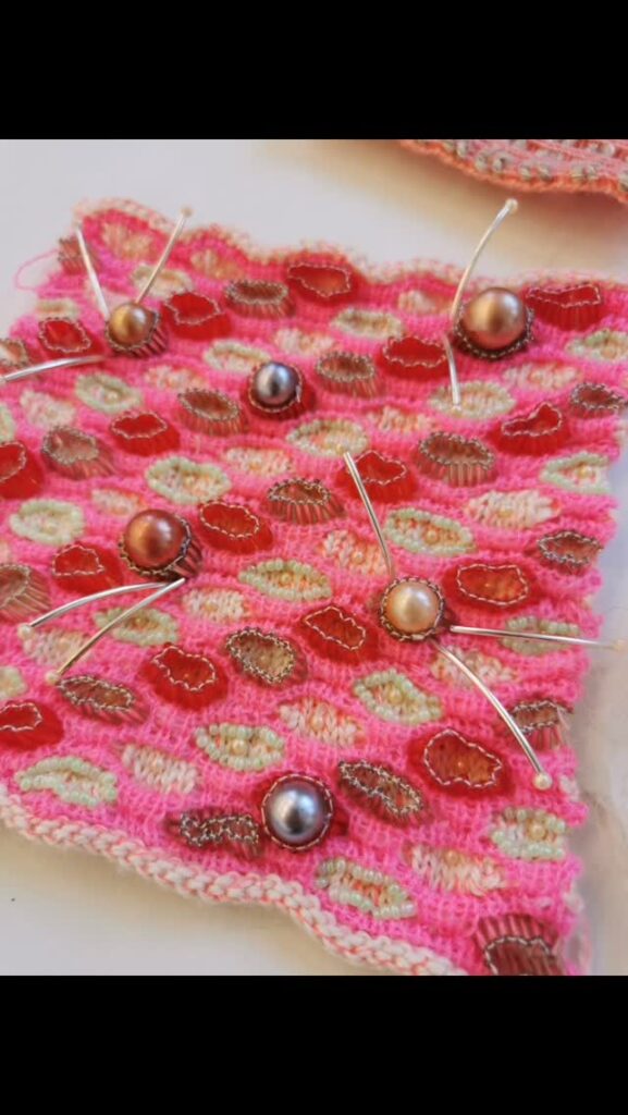 A piece of pink, red, and beige patterned knit fabric is pinned with sewing pins and decorated with several round, metallic beads.