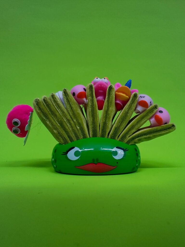 A green, oval-shaped container with a face holds several yellow brushes and colorful ball-shaped objects, each with eyes and faces, against a bright green background.