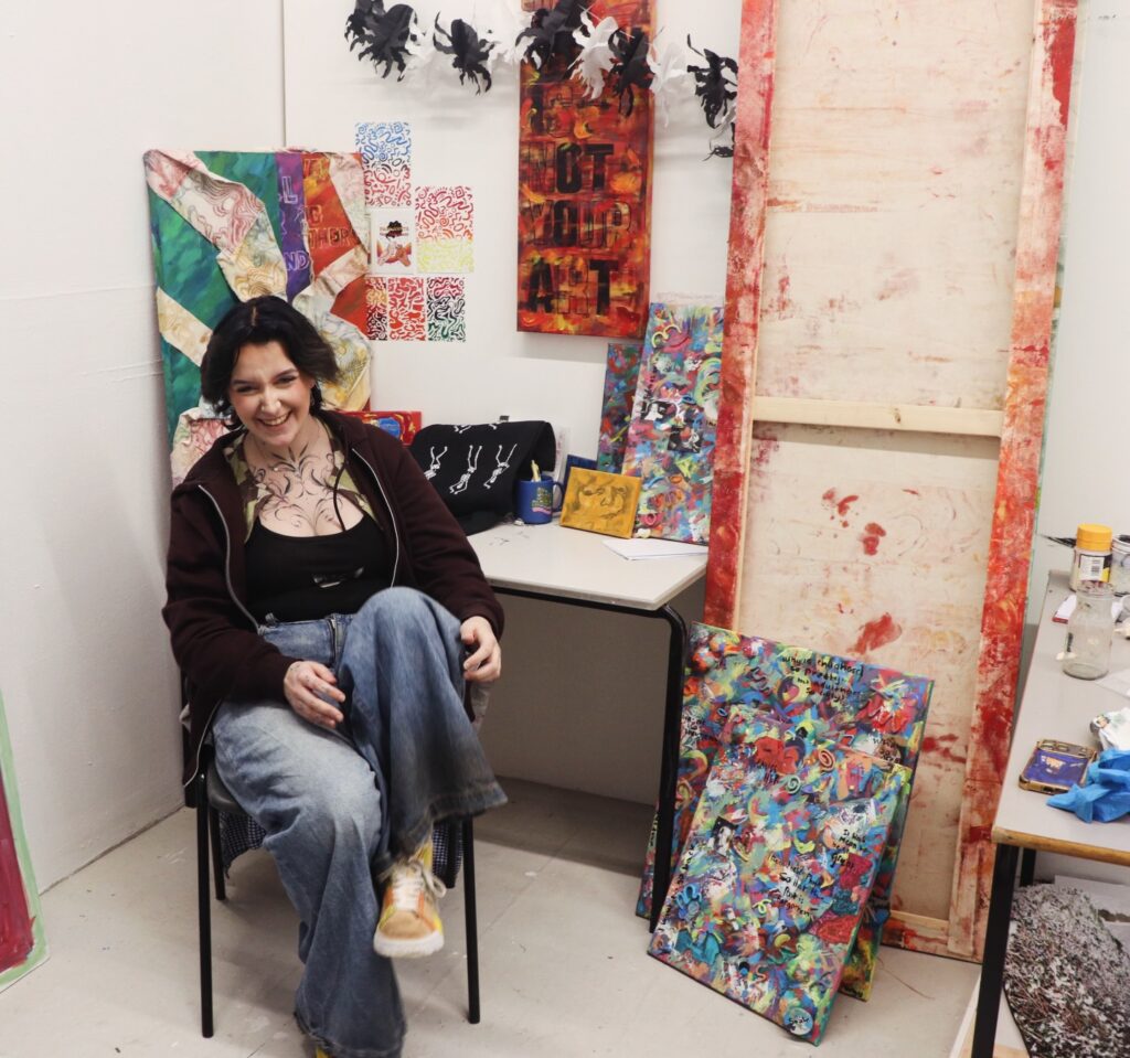 A woman with dark hair, wearing a black top and jeans, sits smiling in an art studio surrounded by vibrant abstract paintings and art supplies. The walls display colorful artworks and creative materials.