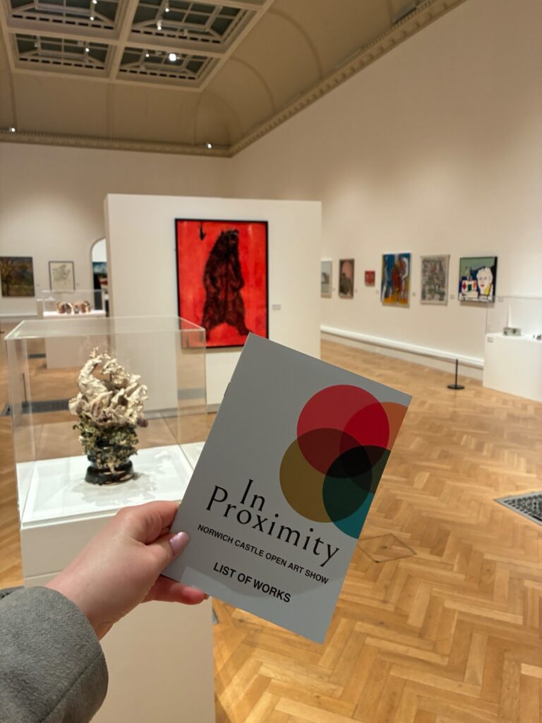 A person holds a brochure titled "In Proximity: Norwich Castle Open Art Show, List of Works" inside an art gallery. Various artworks are displayed on white walls and pedestals around the spacious, well-lit room.