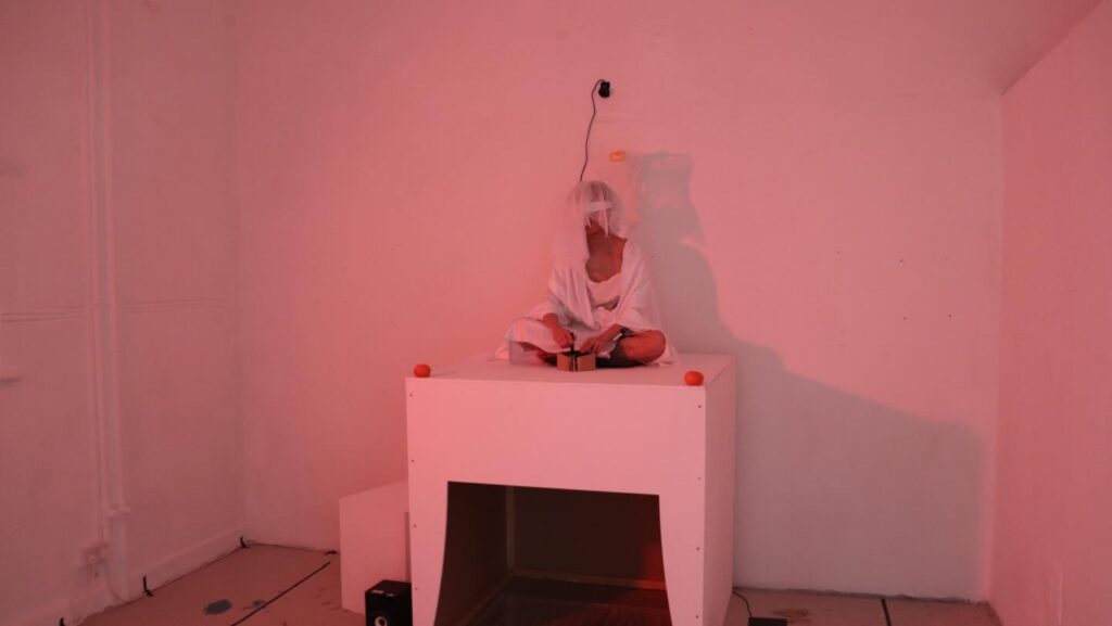 A person in white clothing and a veil sits cross-legged on a white box in a dimly lit, pinkish room. An object is attached to the wall above them, and small items are placed on the box. The scene has an abstract, surreal feel.