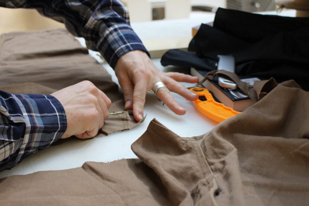 A person in a plaid shirt uses a seam ripper to remove stitches from brown fabric on a worktable, with sewing tools and garments nearby.