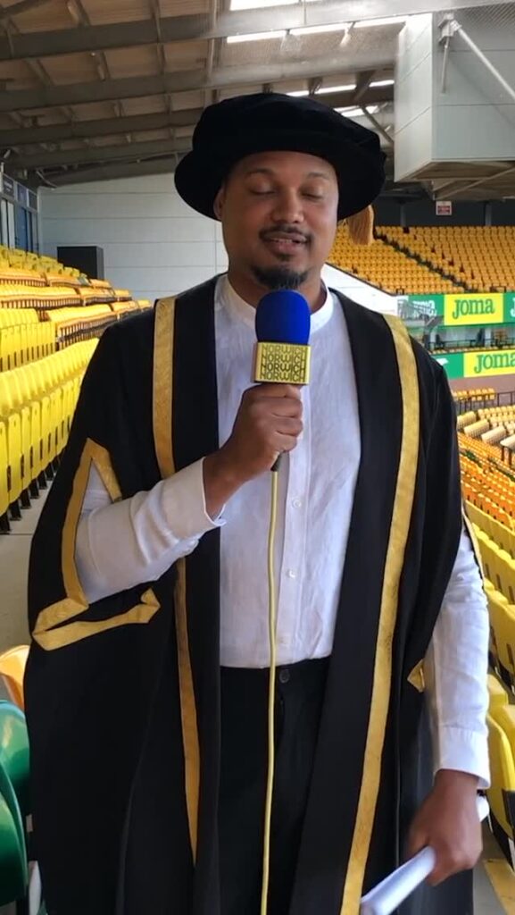 A person wearing academic regalia and holding a blue and yellow microphone stands in a stadium with yellow seats, possibly making an announcement or giving a speech.